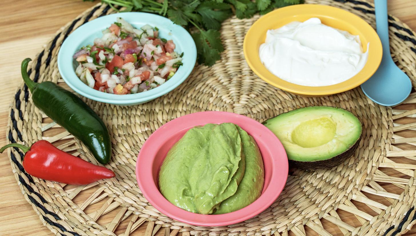 Fresh pico de gallo, guacamole and crema in brightly coloured bowls on a woven placemat